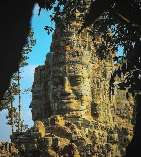 Describe the specific visual elements (smiling stone face, tree framing) while using high-volume travel keywords for Siem Reap.