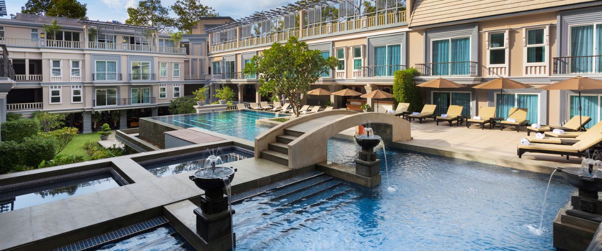 Luxury outdoor swimming pool area at J7 Angkor Hotel in Siem Reap with sun loungers, stone bridges, and balconies.