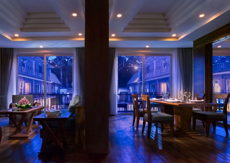 Luxury hotel suite at J7 Angkor Hotel at dusk, featuring an elegant wooden dining table setup, a comfortable living area, and large glass sliding doors opening to a balcony view.