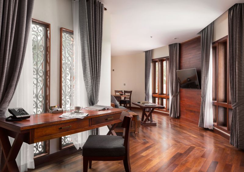 Spacious executive suite living room at J7 Angkor Hotel featuring a wooden writing desk, hardwood floors, a wall-mounted flat-screen TV, and decorative window screens.