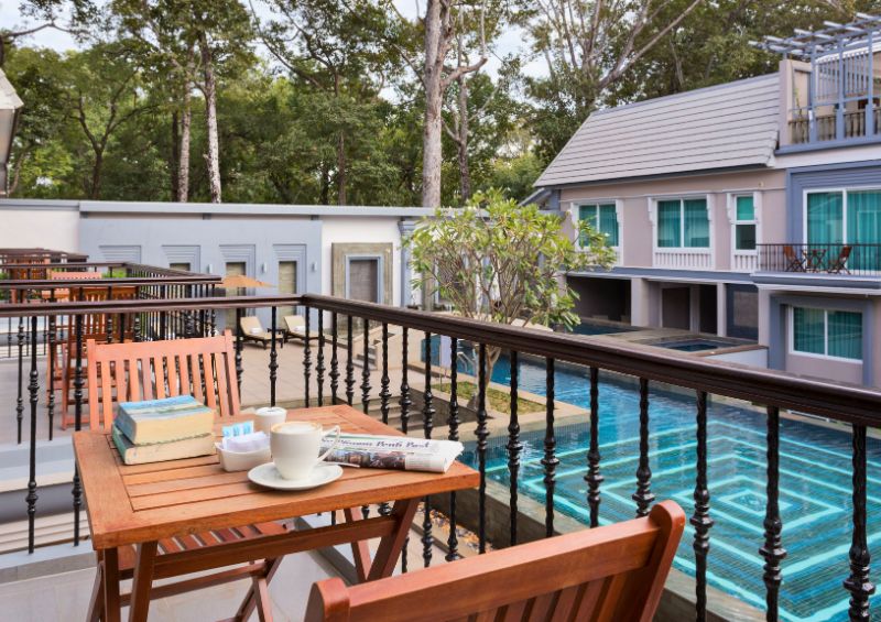 Private room balcony with a wooden table, coffee, and newspaper overlooking the outdoor swimming pool at J7 Angkor Hotel in Siem Reap.