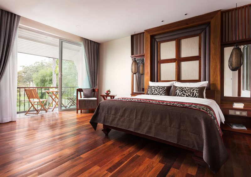 Junior suite hotel room featuring a king bed with an ornamental wooden headboard, polished hardwood floors, and a private balcony with outdoor seating.